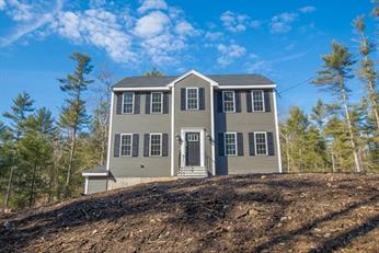 Two-story gray house with black shutters on a hill, surrounded by trees under blue sky.