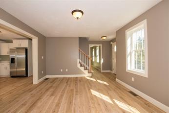 Bright, empty room with wood floors, gray walls, and large windows near a kitchen area.