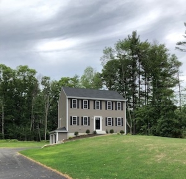 Two-story gray house with black shutters on a grassy hill, trees and cloudy sky behind.
