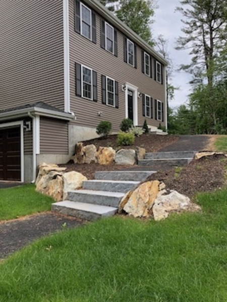 Stone steps with large rocks lead up to a brown house with black shutters and white trim.