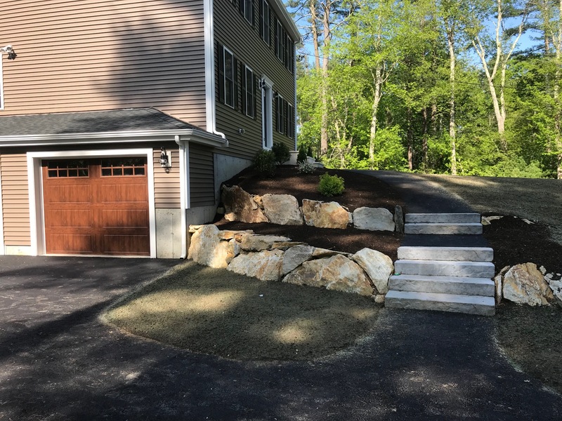 Stone steps and large rocks lead up a landscaped slope next to a house and garage.