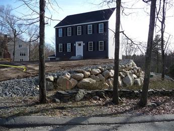 Two-story dark house with white trim, stone retaining wall, bare yard, and leafless trees.