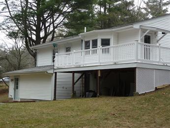 White two-story house with a balcony and lattice skirting, set on a grassy slope.