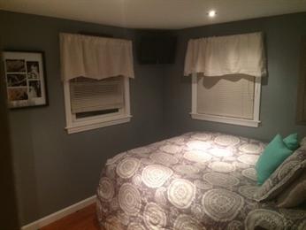 Small bedroom with a patterned bedspread, two windows with blinds, and framed art on the wall.