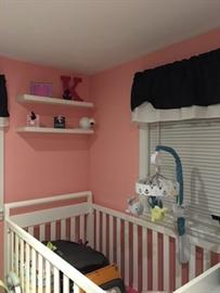 White crib in a pink nursery with shelves, toys, and a window with a black valance.