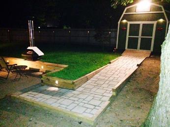 A lit stone path leads to a shed, with a grassy area and chairs on the left, at night.