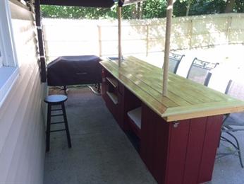 Outdoor bar with a wooden countertop, red base, stool, and patio chairs nearby.