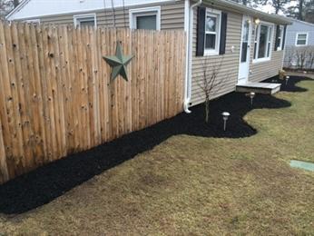 Mulched garden bed along a wooden fence and house, with small plants and lawn lights.