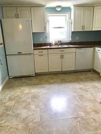 A kitchen with white cabinets, a white fridge, dishwasher, and a window over the sink.