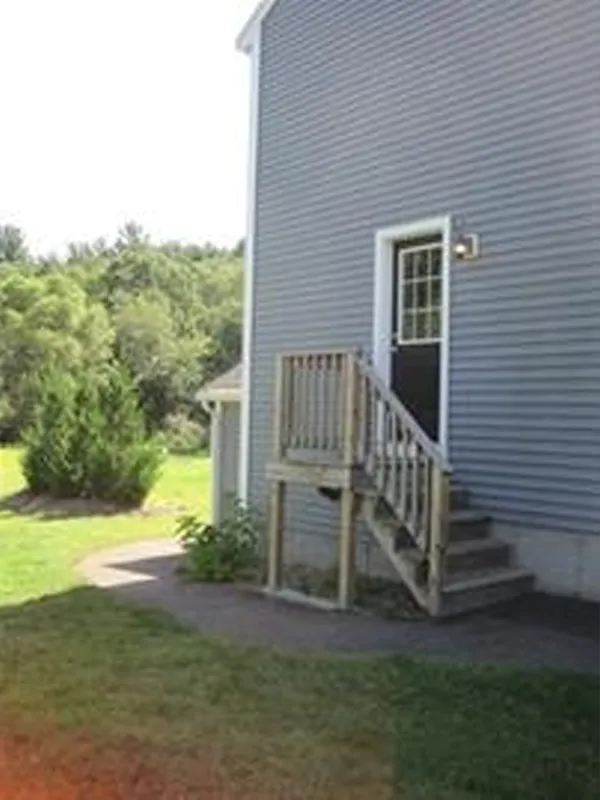 Small wooden stairs lead to a raised door on a blue-gray house, with grass and trees nearby.