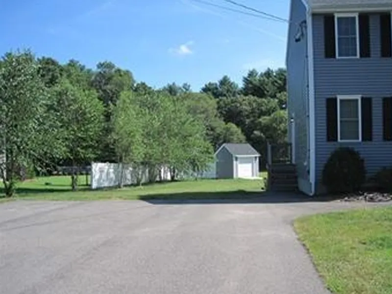 A paved driveway leads to a yard with a shed and trees beside a gray house.