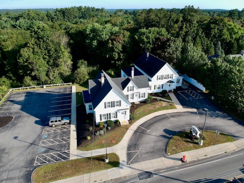 Aerial view of a white building with parking lot, surrounded by trees and greenery.