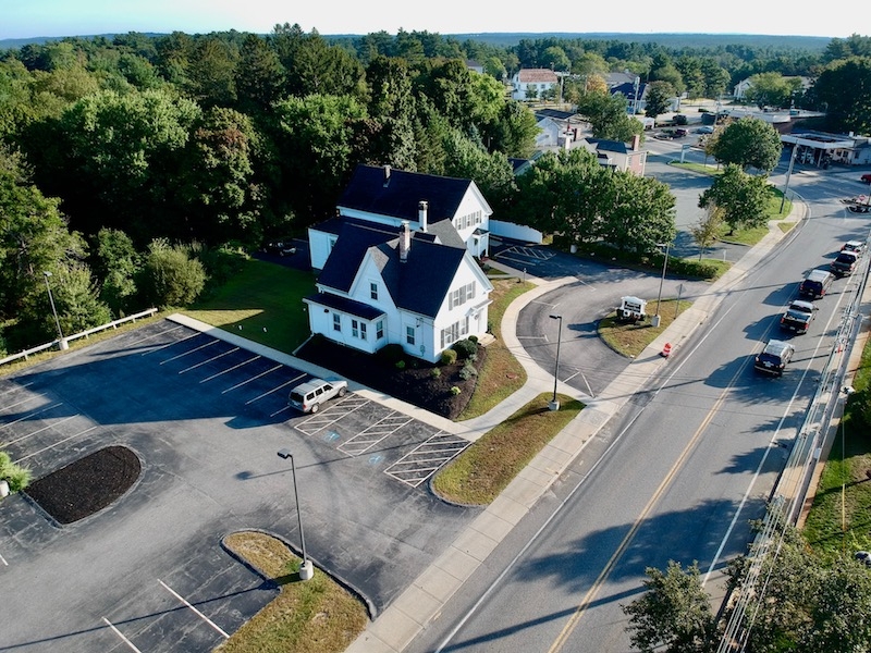 Aerial view of a white house with dark roof by a road, parking lot, and trees.