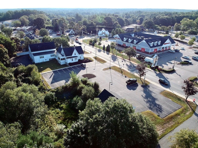 Aerial view of a small commercial area with buildings, parking lots, and surrounding trees.