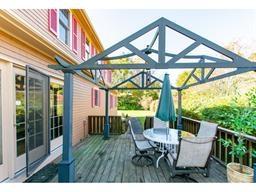 Outdoor deck with a table, chairs, umbrella, and a gazebo frame beside a house.