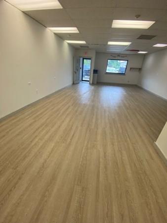 Empty room with wood flooring, white walls, ceiling lights, and windows at the far end.