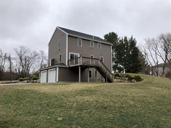 Two-story gray house with a wooden deck and stairs, set on a grassy, sloped yard.