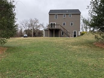 Three-story brown house with deck, surrounded by grass and trees, under a cloudy sky.
