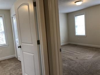 Open doors revealing two carpeted rooms with windows in a neutral-toned interior hallway.
