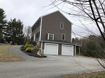 Two-story brown house with double garage, sloped driveway, and trees in the background.