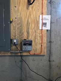 A control box and outlet mounted on plywood on a basement wall with visible wiring.