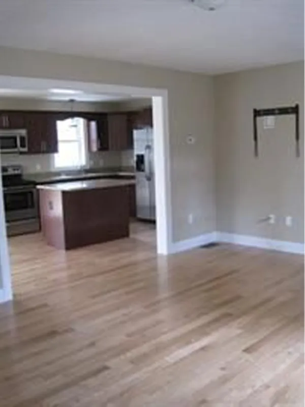 Empty living room with light wood floor, view of kitchen with island and brown cabinets.