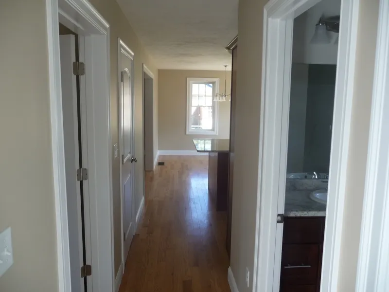 Hallway with light wood floors, beige walls, and doorways; sunlight from window ahead.