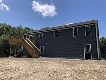 Dark gray house with wooden stairs and deck, set on bare ground under a blue sky.