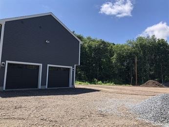 Dark gray garage with two doors, gravel driveway, and trees in the background under blue sky.
