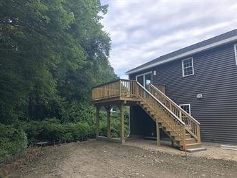 A wooden deck with stairs attached to a dark house, overlooking a dirt backyard and trees.