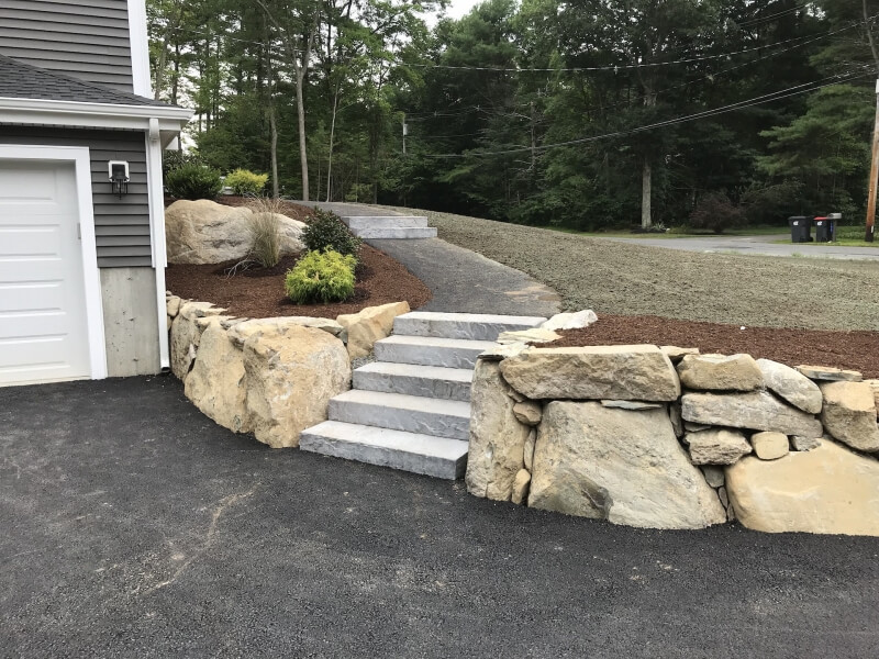 Stone steps with large rock retaining walls lead from a driveway up to a sloped yard.