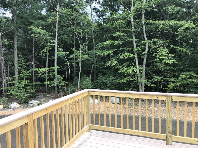 Wooden deck with railing overlooking a paved path and dense green trees in the background.