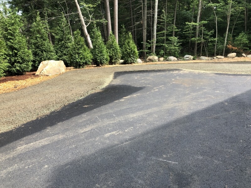 A newly paved driveway curves beside trees and shrubs with rocks along the edge.