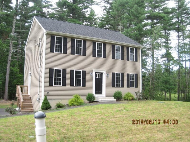 Two-story beige house with black shutters, front steps, and a grassy lawn, surrounded by trees.