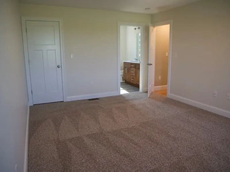 Empty bedroom with beige carpet, white walls, and an open door leading to a bathroom.