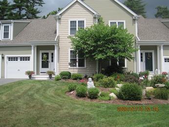 Two-story beige house with a garage, front garden, and green lawn under cloudy sky.