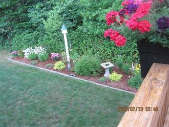 Flower garden with birdbath, lamp post, and red flowers in planter, near green lawn.