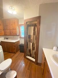 Bathroom with wood floors, small shower with a curtain, sink, and wooden cabinets.