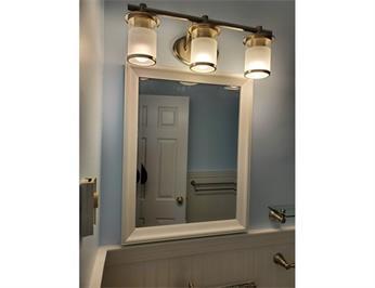 A bathroom mirror with three-light vanity fixture above, reflecting a door and towel rack.