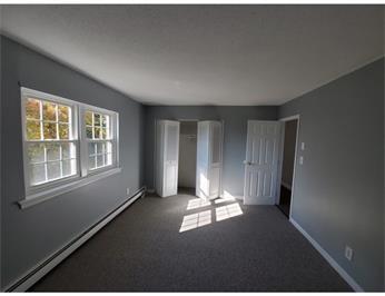 Empty gray-walled room with two windows, closet, and open door, sunlight on the floor.