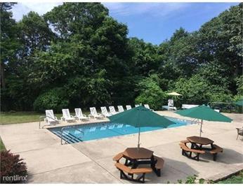 Outdoor pool area with lounge chairs, picnic tables, and green umbrellas surrounded by trees.