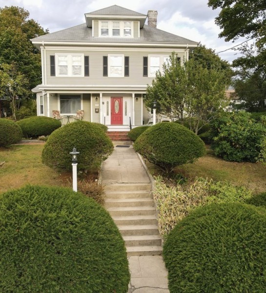 Large two-story house with a red door, trimmed bushes, and concrete steps leading up.