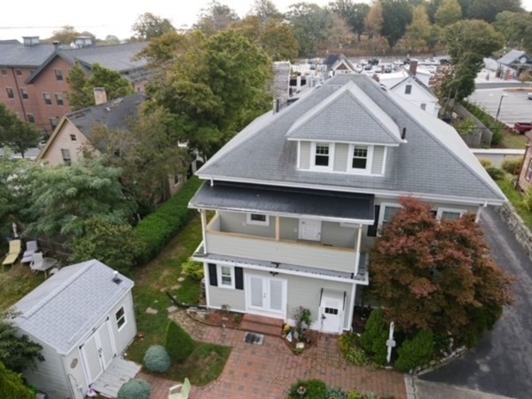 Aerial view of a two-story gray house with a porch, shed, and surrounding trees.