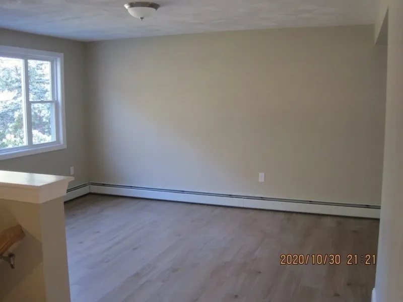 Empty room with light wood floor, beige walls, and a large window letting in sunlight.