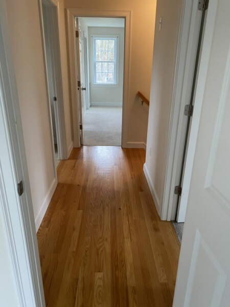 A hallway with wooden floors leading to a bright room with a window at the end.