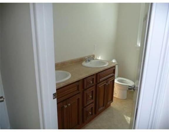 Bathroom with double sinks, wooden vanity, and a toilet in the corner near a window.