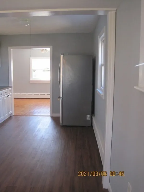 A silver refrigerator blocks a window in a gray-walled kitchen with wood flooring.