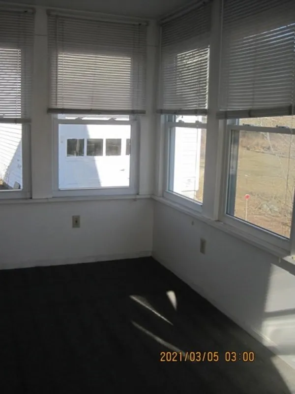 Small sunroom with several windows, blinds, white walls, and dark flooring, sunlight coming in.
