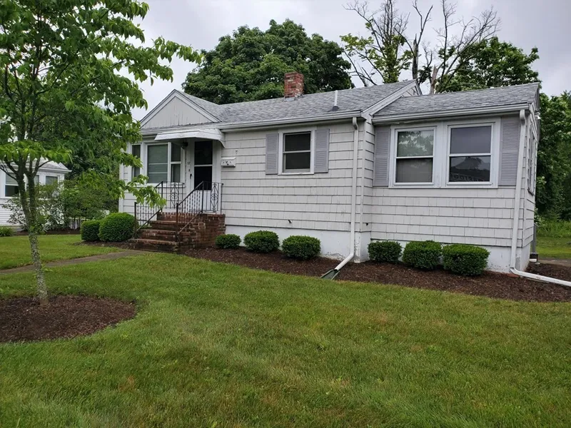Small gray house with white trim, front steps, shrubs, and a green lawn on a cloudy day.