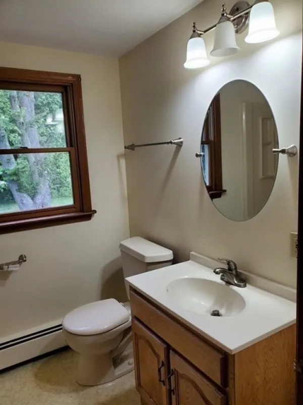 A simple bathroom with a toilet, sink, oval mirror, and window with outdoor view.
